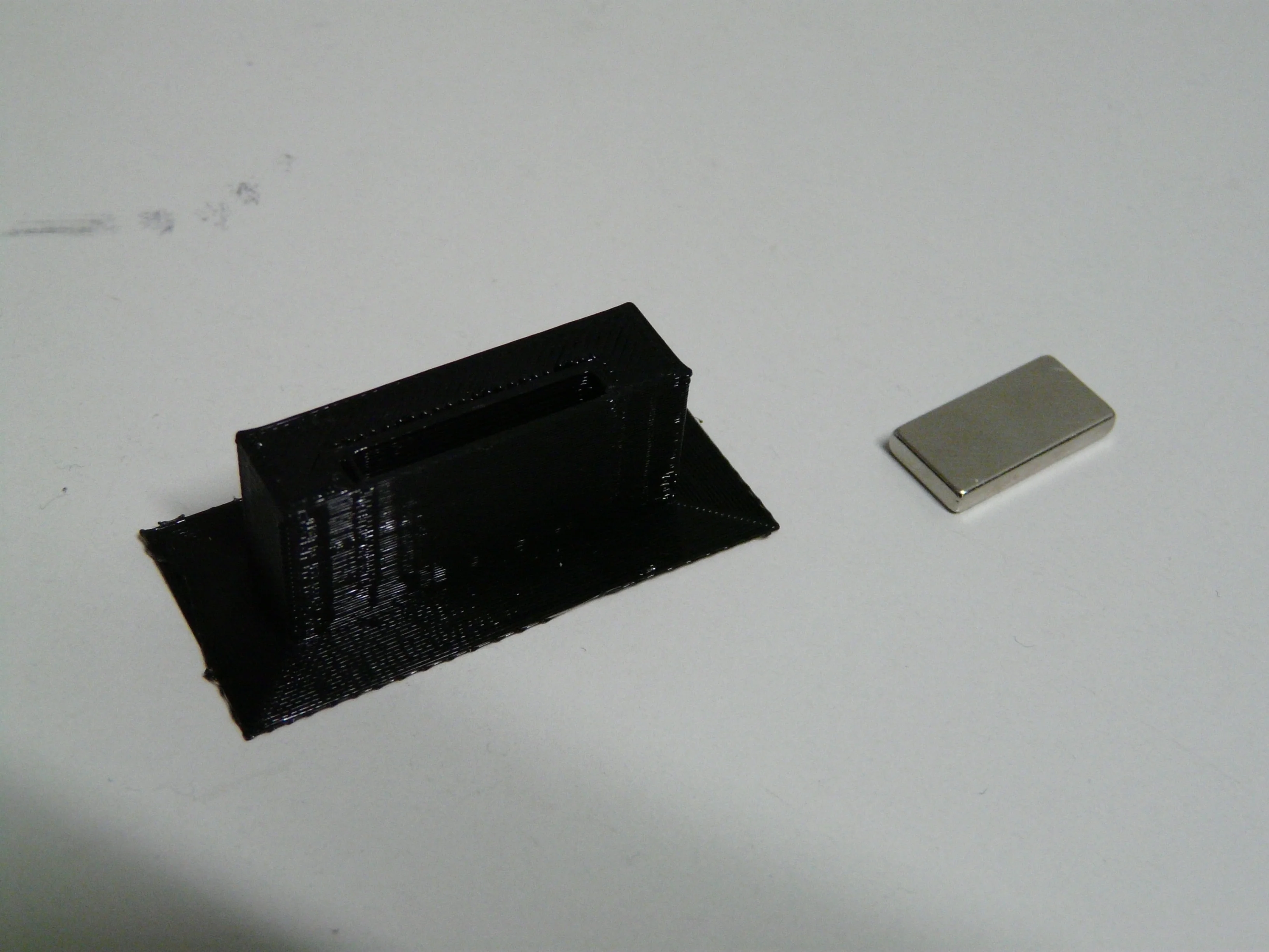 Musta 3D-tulostettu ovenpidike ja erillinen suorakulmainen magneetti.
